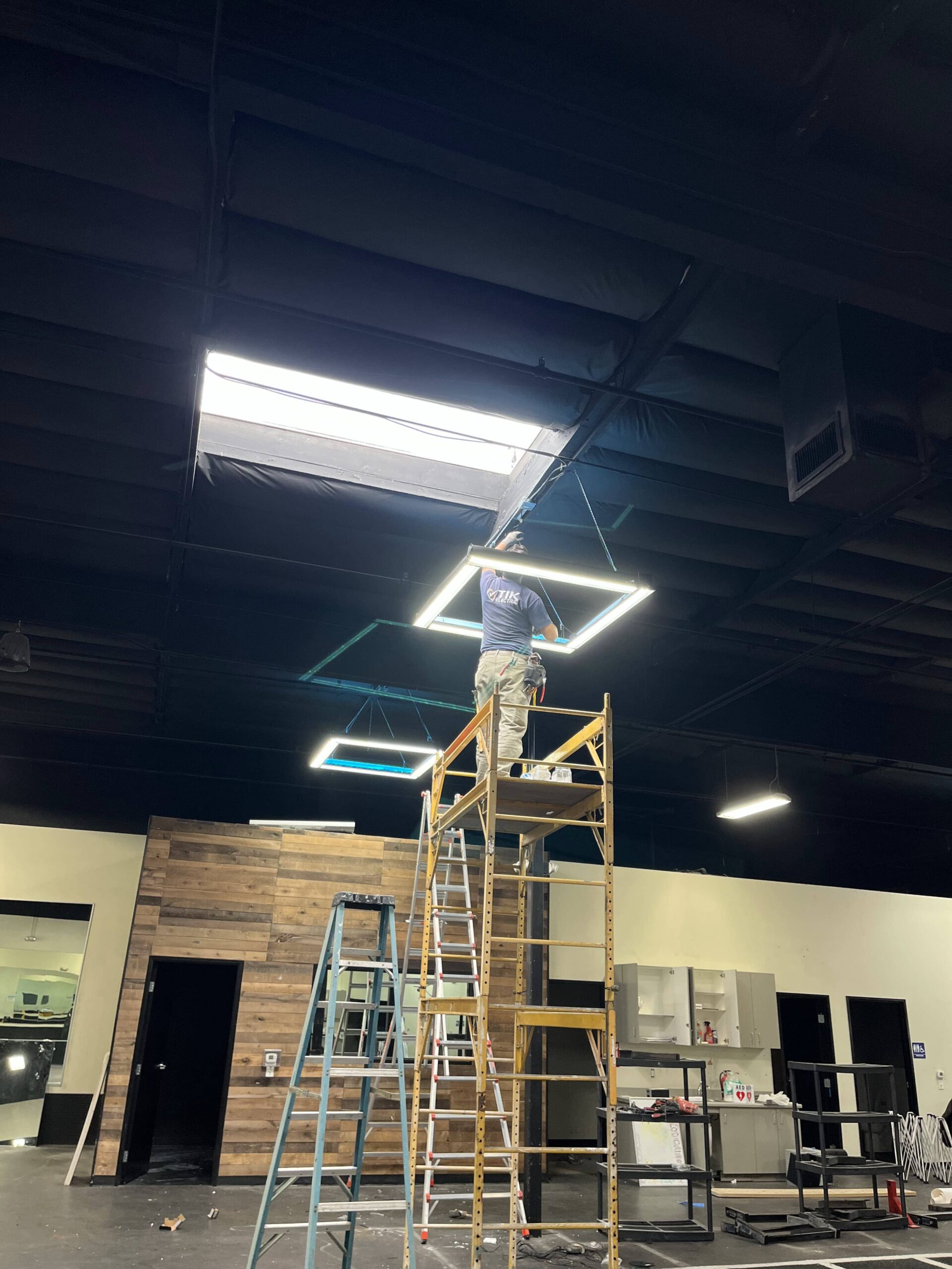 Tech on scaffold installing ceiling LED grid lights in showroom — neat routing and aligned panels